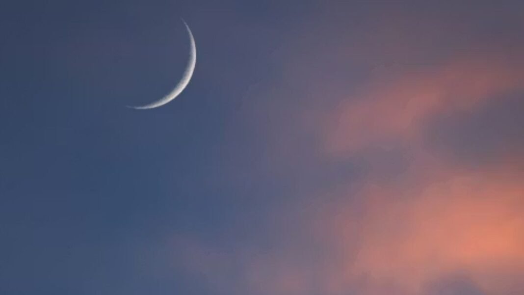 Thin crescent moon visible in evening sky with soft pink clouds