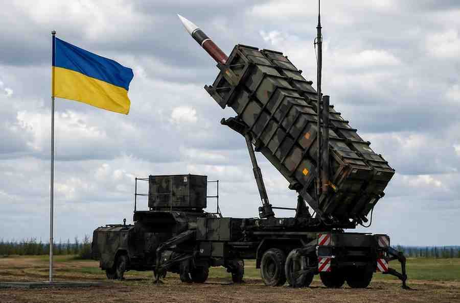 Patriot missile launcher stands beside a Ukrainian flag under cloudy skies.